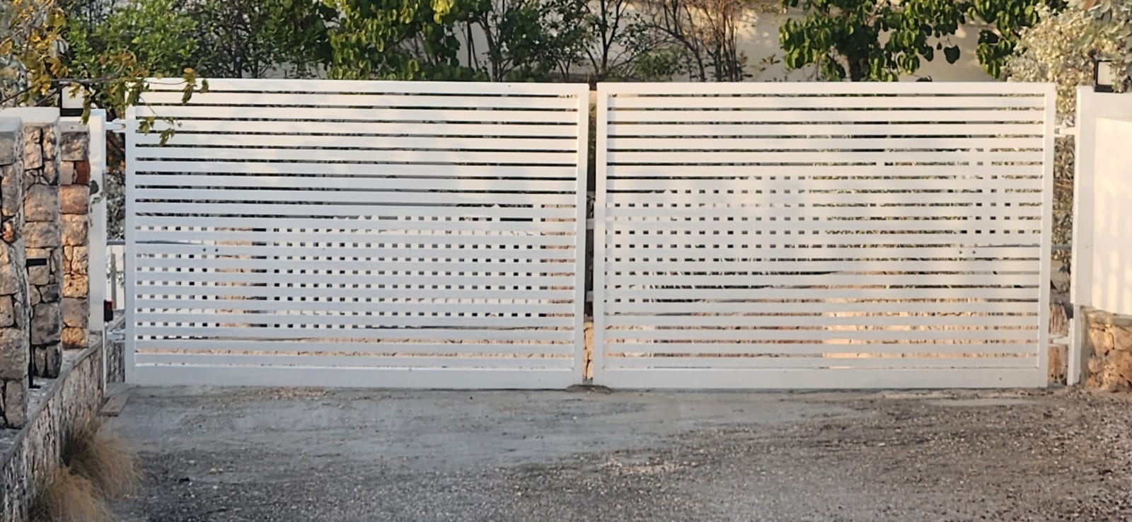 White horizontal slat aluminium driveway gate set between stone pillars in Anguilla